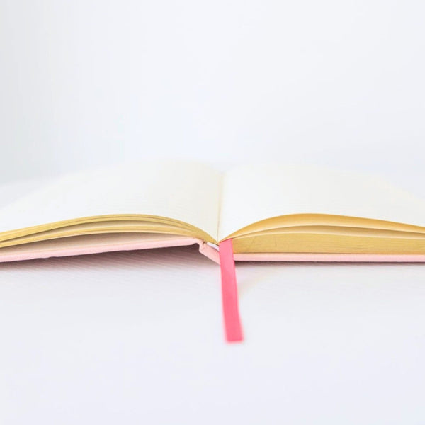 An open notebook with cream-colored pages, yellow-gold inner edges, a pale pink cover, and a bright pink ribbon bookmark, set against a clean white background.