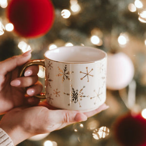 Person holding a mug with gold snowflake patterns against a festive background with lights and decorations.