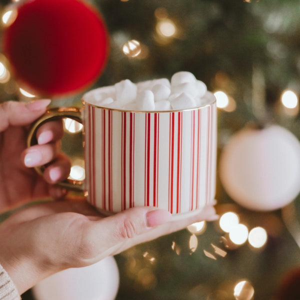 Hand holding a mug with red and white stripes filled with marshmallows, Christmas tree in the background