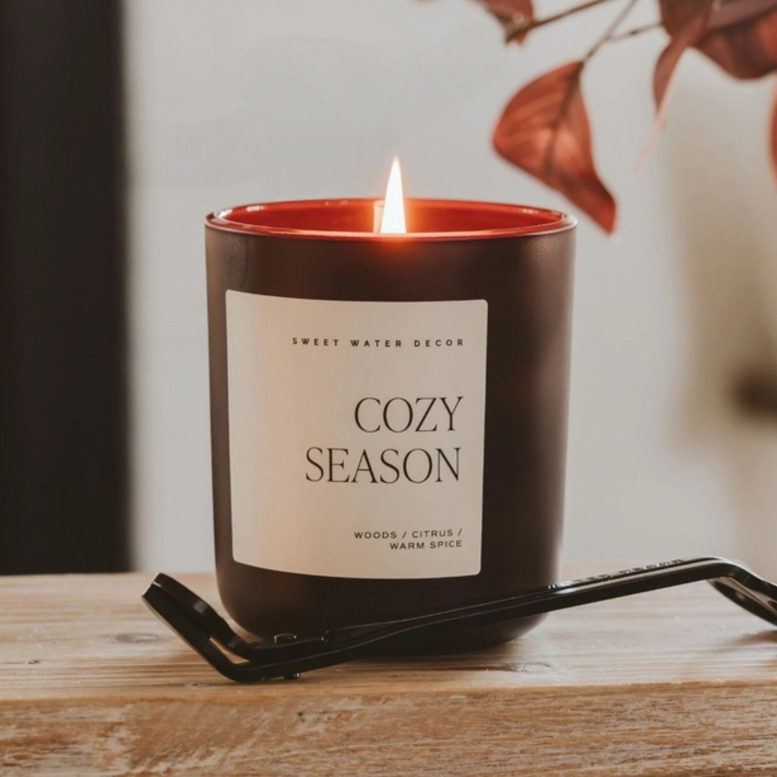 A lit candle in a dark brown glass jar with a cream label reading 'COZY SEASON' and 'SWEET WATER DECOR'. The candle sits on a wooden surface with a black metal candle trimmer and fall leaves in the background | Build A Luxury Custom Gift Box with Luxe & Bloom