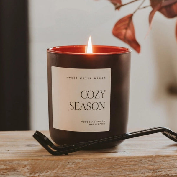 A lit candle in a dark brown glass jar with a cream label reading 'COZY SEASON' and 'SWEET WATER DECOR'. The candle sits on a wooden surface with a black metal candle trimmer and fall leaves in the background | Build A Luxury Custom Gift Box with Luxe & Bloom