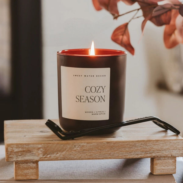 A lit candle in a dark brown glass jar with a cream label reading 'COZY SEASON' and 'SWEET WATER DECOR'. The candle sits on a wooden tray with a black metal candle trimmer and fall leaves with a blurred background | Build A Luxury Custom Gift Box with Luxe & Bloom