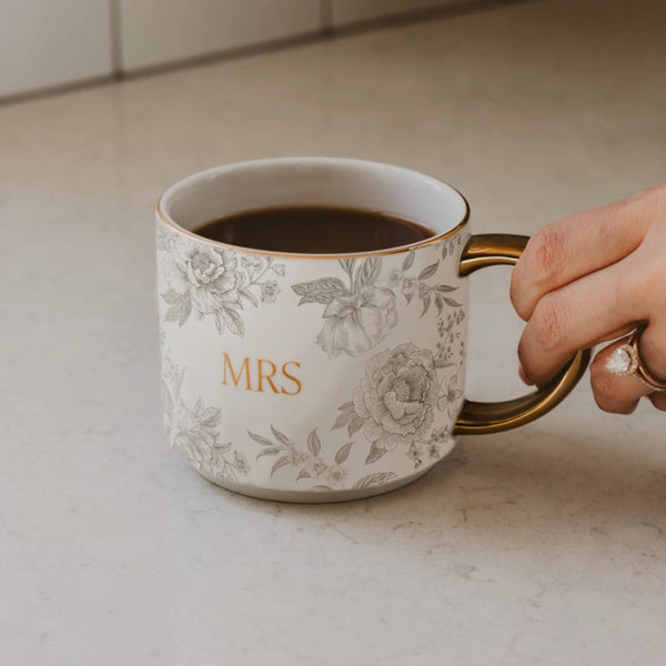 Sweet Water Decor Mug with floral design and 'Mrs.' text, held by a hand on a light surface.