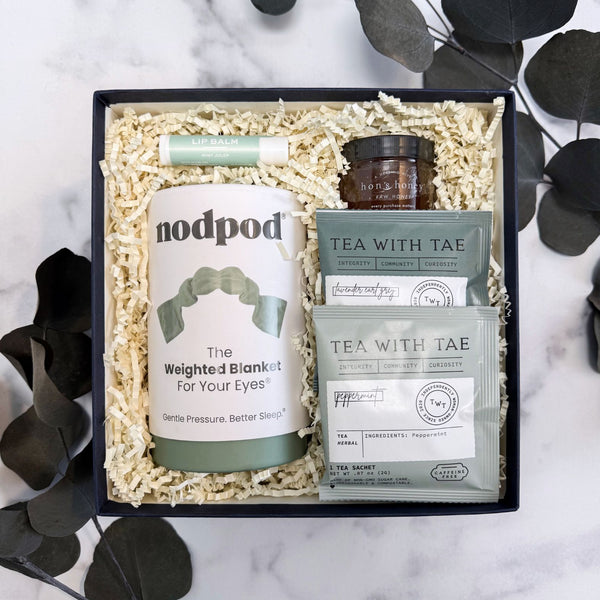 Well Wishes Luxury Gift Box set containing a sage green nodpod Weighted eye mask, two green Tea With Tae tea packets, a small jar of honey, and a green lip balm tube, arranged in crinkle paper inside a navy gift box on a marble surface with eucalyptus leaves around it.