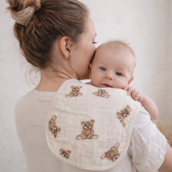 Woman holding a baby with Ali + Oli teddy bear bamboo muslin burp cloth over her shoulder against a plain background