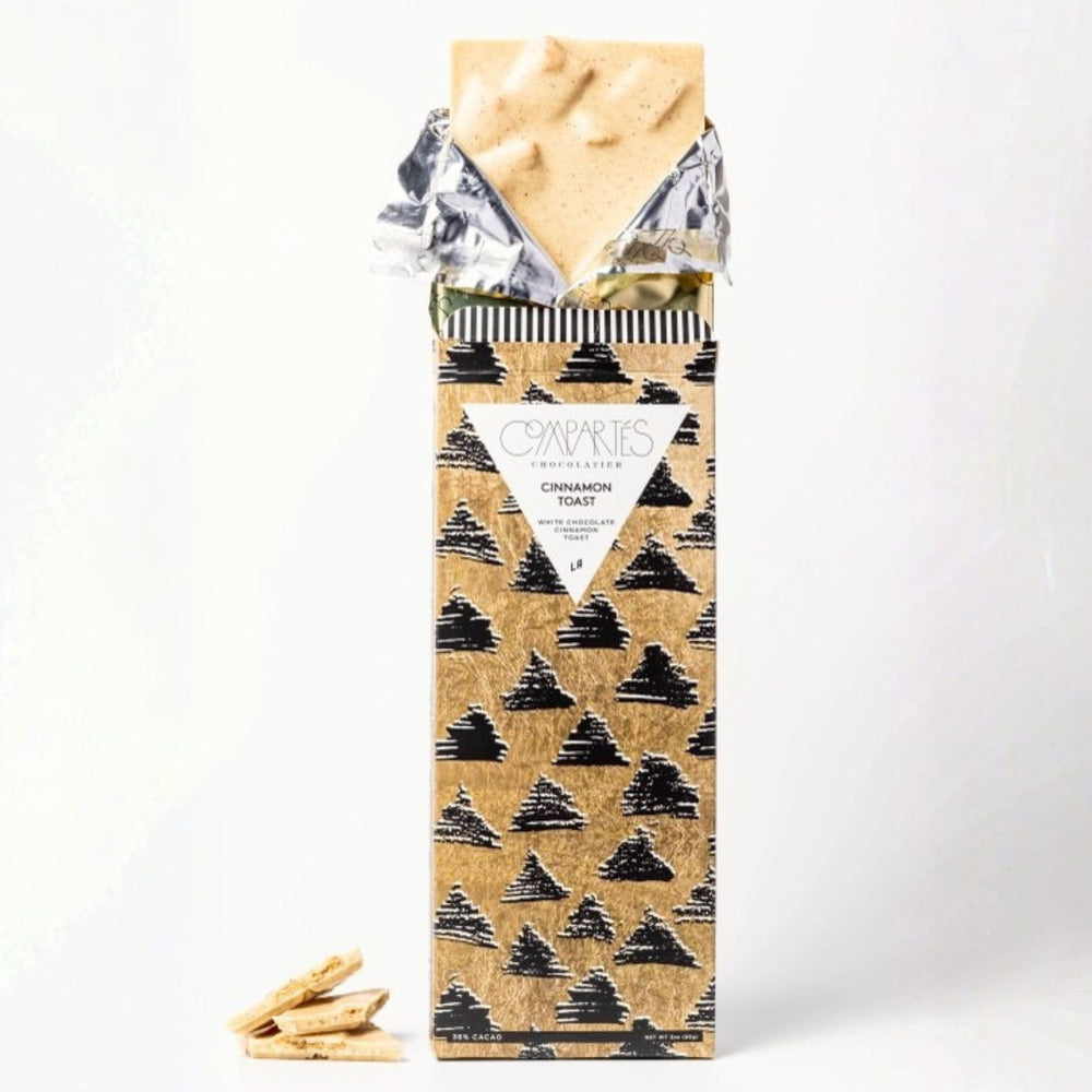 A partially unwrapped gourmet chocolate bar from the brand Compartés Chocolate, featuring a patterned gold wrapper with black diamond silhouettes and a white label reading 'Cinnamon Toast'. The bar itself is pale cream-colored.