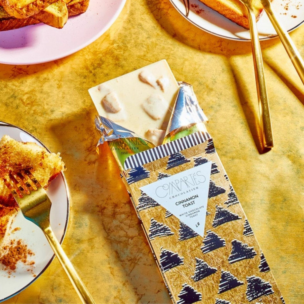 A partially unwrapped gourmet chocolate bar from the brand Compartés Chocolate, featuring a patterned gold wrapper with black diamond silhouettes and a white label reading 'Cinnamon Toast'. The bar itself is pale cream-colored.