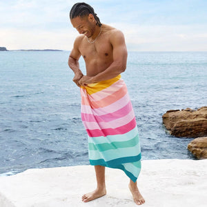 Man standing on a rocky beach wearing a colorful striped towel. Dock & Bay Coastal Candy Quick Dry Towel | Build A Luxury Custom Gift Box with Luxe & Bloom