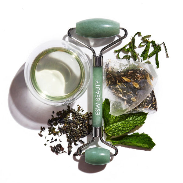 Green ESW Beauty Jade roller with jar of cream, tea leaves, and mint leaves on a white background