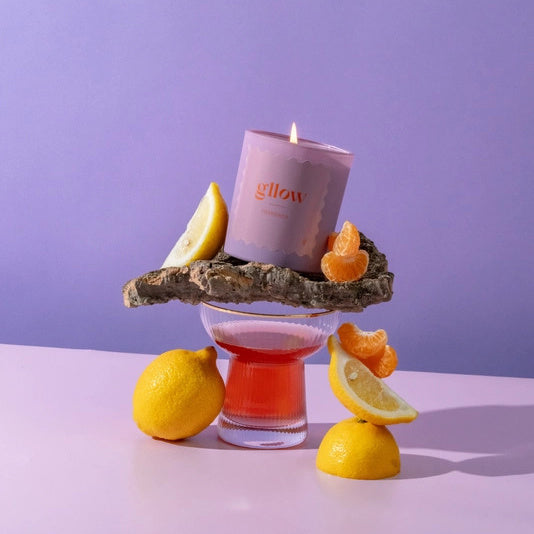 Gllow Cherished Scented Candle on a decorative stand with lemons and oranges against a purple background | Build A Luxury Custom Gift Box for Women with Luxe & Bloom