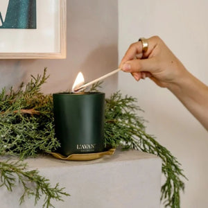 A dark green L'AVANT Winter Fir candle in a gold holder, lit with a wooden match held by a person's hand, styled with green pine branches on a light gray fireplace mantle.
