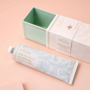 Lollia Snowdrop hand cream tube and its matching box on a soft peach background. The tube is white with pale blue floral patterns and gold lettering, and the box is white with a mint green interior.