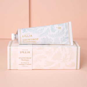 A tube of Lollia Snowdrop Hand Cream resting on top of its matching decorative box, set against a soft peach-pink background.