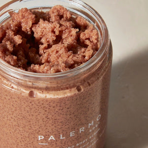 An open clear jar filled with a pink granular scrub, with the brand name PALERMO visible on the front label, set against a neutral beige background. Palermo Body Revitalizing Body Scrub | Build A Luxury Custom Gift Box for Women with Luxe & Bloom