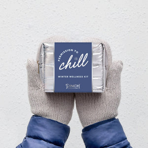 Pair of mittens holding a Pinch Provisions Winter Wellness Kit labeled 'Permission to Chill' on a light gray background.