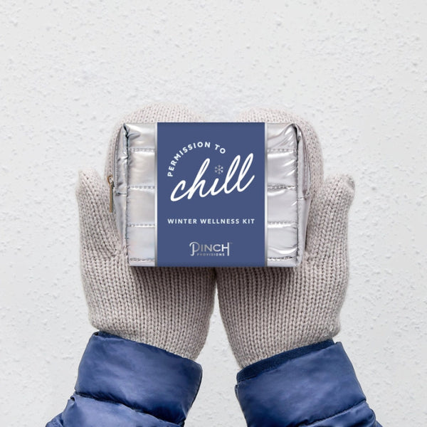 Pair of mittens holding a Pinch Provisions Winter Wellness Kit labeled 'Permission to Chill' on a light gray background.