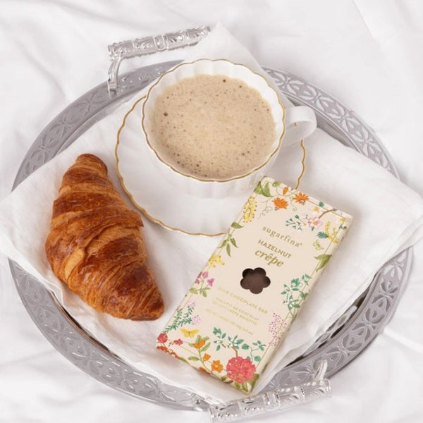 Cup of coffee, croissant, and a Sugarfina Hazelnut Crèpe Chocolate Bar package with floral design on a silver tray with a white background