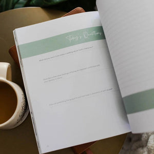 Open The Daily Grace Co. Worthy: embracing your identity in Christ Devotional with a green stripe titled 'Today's Questions' next to a mug on a wooden surface.
