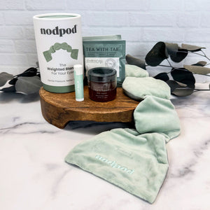 A styled product arrangement on a marble surface with a wooden tray, featuring a white and sage green nodpod container with the weighted eye mask laid out on the surface, a small jar of honey, two tea packets, and a Mint Julip lip balm tube. The background is a white brick wall with decorative eucalyptus leaves.